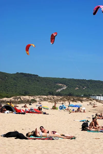 Turistas tomando el sol en la playa con cometas de surf contra el cielo azul, Cabo Trafalgar, España . — Foto de Stock