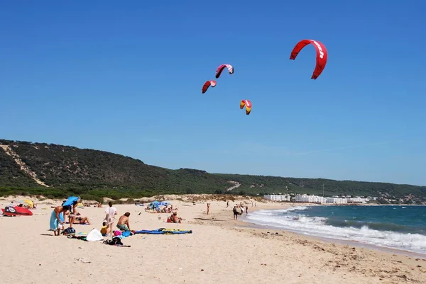 Turistas tomando el sol en la playa con cometas de surf contra el cielo azul, Cabo Trafalgar, España . — Foto de Stock