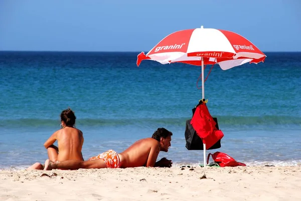 Parejas jóvenes con una sombrilla tomando el sol junto al borde de las aguas en la playa con vistas al mar, Zahara de los Atunes, España . — Foto de Stock