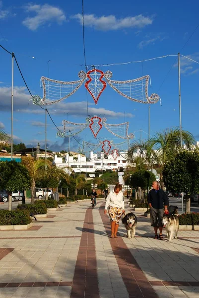 Świąteczna dekoracja uliczna w centrum miasta z parą spacerów swoich psów na pierwszym planie w ciągu dnia, La Cala de Mijas, Hiszpania. — Zdjęcie stockowe