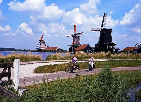 前景でサイクリングをするカップルと川のほとりに沿って風車の列, ザンセ・シャンズ, オランダ. — ストック写真