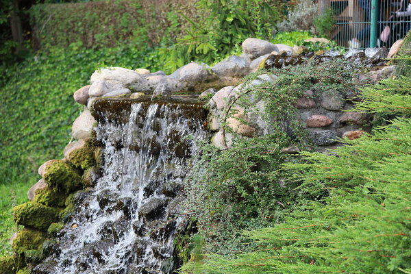 The photo shows an artificial waterfall in Gdansk Zoo, surrounded by greenery and a small pond.