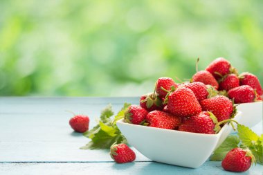 Tahta masada beyaz kaselerde taze çilek, yeşil doğa arka planı.