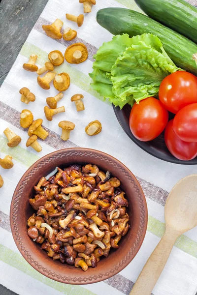 Gebratene Pfifferlinge mit Zwiebeln in rustikaler Schüssel und Teller mit frischem Gemüse für Salat im Hintergrund — Stockfoto