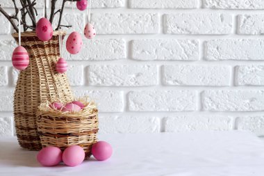 Hasır bir vazoda süslü ağaç dalları ve beyaz arka planda hasır sepette pembe renkli yumurtalar olan Paskalya kompozisyonu. Boşluğu kopyala