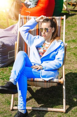 Festivalde dinlenin. Kız katlanır karyolada oturuyor ve uzaklığa bakıyor. Güneş gözlüğü. Elbiselerin mavi rengi. güneşli bir gün