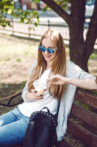 Sarışın gülümsüyor. Sokakta bir bankta oturuyor. Güneşli bir gün. Güneş gözlüğü. Pipetle canlandırıcı kokteyl.