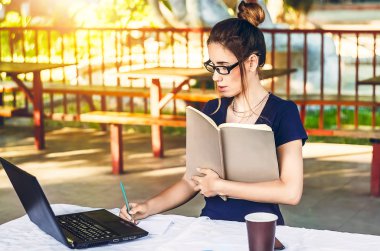 Kız bir dizüstü bilgisayar için temiz havada çalışıyor. Elinde notlar için bir defter tutuyor. Bir bardak sıcak içecek. Koruyucu gözlük. Güneşli bir gün. iş, internet, öğrenim.