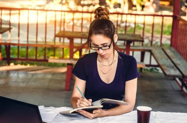 ders çalış. Şeffaf çerçeveli gözlüklü bir kız günlüğüne yazıyor. Laptop masanın üstünde..