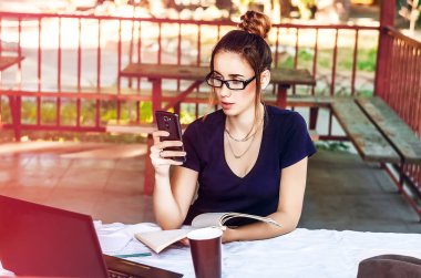 Gözlüklü kız telefonu tıklatıyor. Kablosuz internet, sosyal ağlar, iletişim. dizüstü bilgisayar ve defter