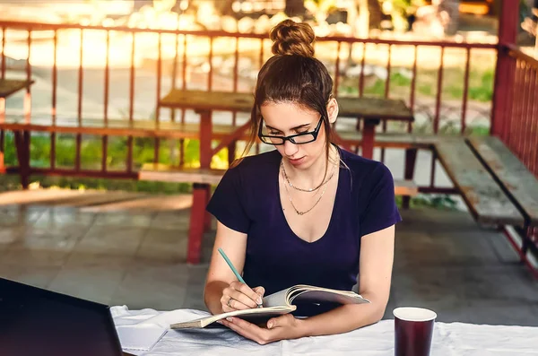 ders çalış. Şeffaf çerçeveli gözlüklü bir kız günlüğüne yazıyor. Laptop masanın üstünde..