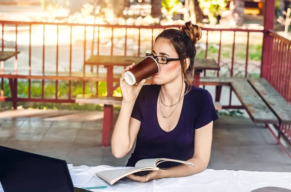 Kız, masada oturmuş, temiz havada kahve içiyor. Laptop ve telefon var. Not defteri. çalış, çalış.