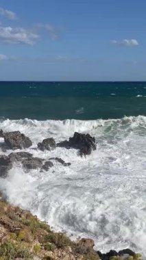 Akdeniz açık güneşli havada köpürüyor, dalgalar güçlü bir şekilde kıyı kayalarına çarpıyor ve su ve köpüğü havaya yolluyor.