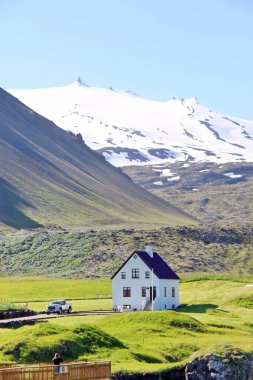 A solitary white house with a black roof stands on lush green grass beneath a dramatic, snow-capped mountain in bright summer daylight, likely in rural Iceland.