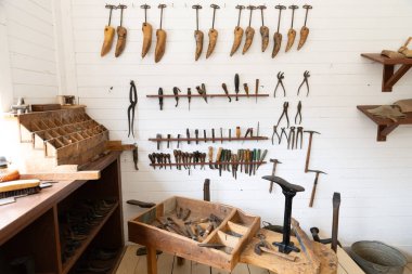 Geleneksel ayakkabı yapma aletleriyle dolu ahşap raflar ve sabitler, beyaz bir duvara güzelce yerleştirilmiş. Ahşap tezgahta çekiç, kerpeten, çan ve örs ayakkabı gibi çeşitli ayakkabı aletleri bulunur.. 