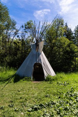 Geleneksel bir kızılderili çadırı güneşli bir günde ağaçlarla çevrili çimenli bir düzlükte duruyor. Uzun tahta direklerle desteklenen tuval yapısı, doğayla uyum içinde yaşayan kültürel mirası ve açık havayı temsil ediyor..