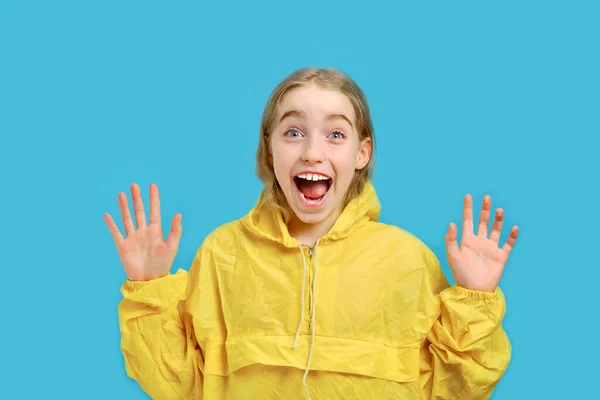 Καυκάσιο χαρούμενο κορίτσι με κίτρινο μπουφάν σήκωσε τις παλάμες της. Η έννοια των συναισθημάτων και των εκφράσεων του προσώπου. Στούντιο απομονωμένο πορτρέτο μισού μήκους σε μπλε φόντο. — Φωτογραφία Αρχείου
