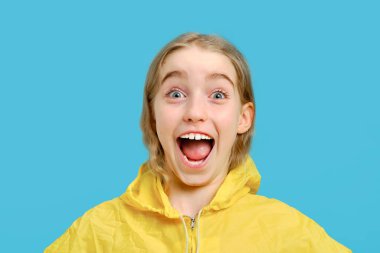 Kafkasyalı, sarı ceketli neşeli küçük kız ağzını açtı. Duygular ve yüz ifadeleri kavramı. Stüdyo mavi bir arkaplanda yarı boy portre izole.