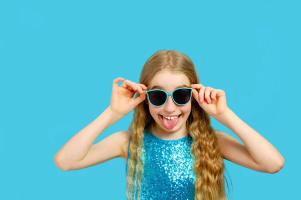 Schöne verschmitzte Kaukasierin mit Sonnenbrille zeigt Zunge. Das Mädchen trägt ein wunderschönes blaues Kleid. Das Konzept von Emotionen und Mimik. — Stockfoto
