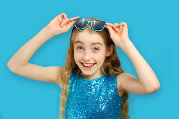 Красивая веселая белая маленькая девочка в солнечных очках и смотрит в камеру. Девушка одета в красивое голубое платье. Концепция эмоций и мимики . — стоковое фото