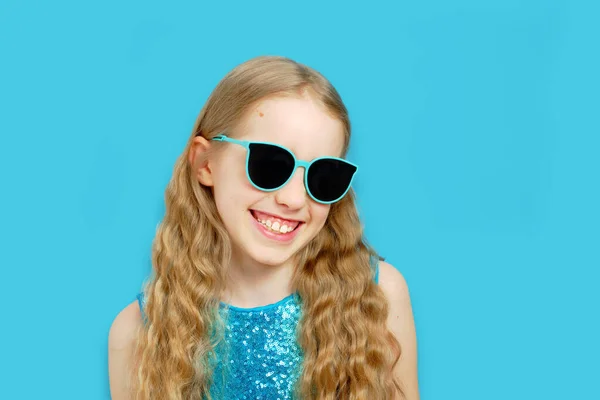 Schöne kleine kaukasische Mädchen mit Sonnenbrille in einem blauen Kleid. Das Mädchen lächelt und blickt in die Kamera. Vereinzeltes halblanges Porträt auf blauem Hintergrund. — Stockfoto