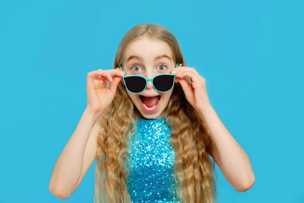 Красивая озорная белая маленькая девочка сняла солнечные очки и смотрит в камеру. Девушка одета в красивое голубое платье. Концепция эмоций и мимики. — стоковое фото