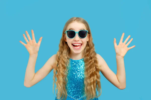 Schöne fröhliche kleine kaukasische Mädchen in einem blauen Kleid blickt in die Kamera. Sie zeigt ihre Hände und freut sich. Vereinzeltes halblanges Porträt auf blauem Hintergrund. — Stockfoto
