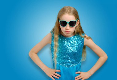 Beautiful Caucasian little girl in sunglasses and blue dress looks angrily into the camera. Emotions and adolescent crisis concept. Isolated half-length portrait on blue background clipart