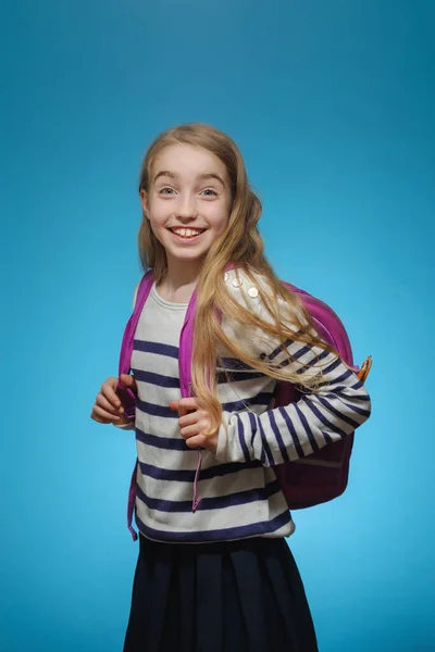Маленькая белая девочка со школьным рюкзаком на голубом фоне изолирована. Девушка смотрит в камеру и улыбается.. — стоковое фото