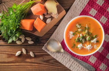  Kırmızı kareli masa örtüsünün üzerindeki ahşap masada peynirli ve çekirdekli balkabağı kremalı çorba. Yakınlarda yemek pişirmek için malzemeler var.