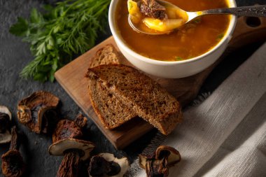 Siyah arka planda beyaz yuvarlak tabakta porcini mantarlı mantar çorbasını kapat. Yakınlarda siyah ekmek ve kurutulmuş porcini mantarı var..