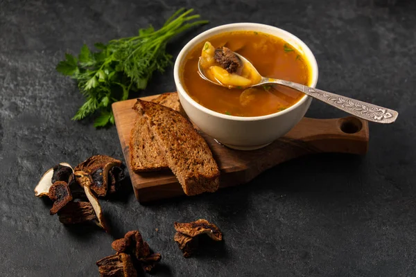 Mushroom soup with porcini mushrooms in a white round plate on a black background. Nearby lies dark bread and dried porcini mushrooms. — Stock Image Mushroom soup with porcini mushrooms in a white round plate on a black background. Nearby lies dark bread and dried porcini mushrooms. Royalty Free Stock Images