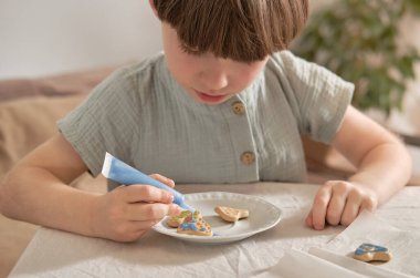 Evde renkli kremalarla kurabiye süsleyen genç bir çocuk. Rahat çocukluk anı, yaratıcı pişirme etkinliği, aile yaşam tarzı ve tatil hazırlama konsepti..