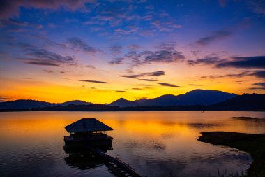 Güneş, önünde büyük bir su kaynağı ve küçük bir evin gölgeleriyle bir dağın arkasında batıyor. Resimde bir burun var..