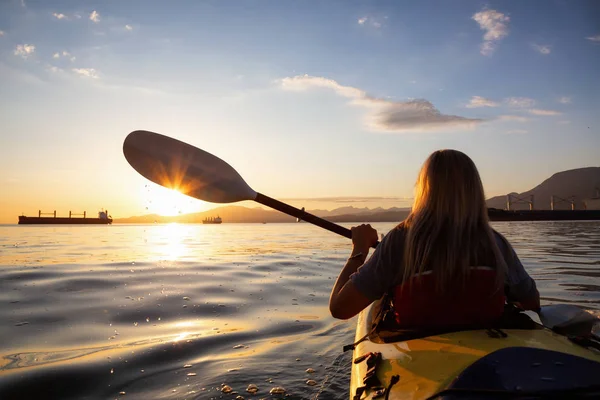 Woman on a kayak is paddeling in the ocean during a vibrant sunset. Taken in Vancouver, British Columbia, Canada. Concept: adventure, holiday, lifestyle, sport, recreation, activity, vacation