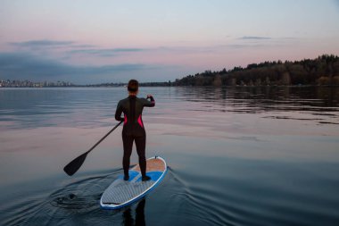 Bir kürek tahtada maceracı kız canlı bir gün batımı sırasında arka planda şehir şehir ile okyanusa paddeling var. Alınan İspanyolca bankalar, Vancouver, British Columbia, Kanada.