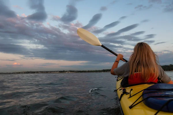 Woman on a kayak is paddeling in the ocean during a vibrant sunset. Taken in Vancouver, British Columbia, Canada. Concept: adventure, holiday, lifestyle, sport, recreation, activity, vacation