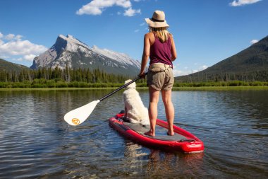 Banff, Alberta, Canada - June 19, 2018:  Adventurous woman on a paddle board with her dog is enjoying the beautiful sunny summer day on the lake. clipart