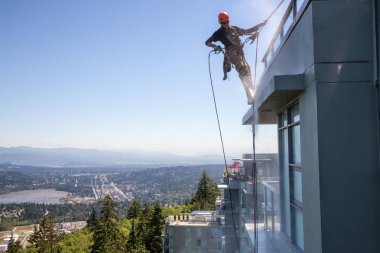 Burnaby Mountain, Vancouver, British Columbia, Canada - July 011, 2018: High rise rope access window cleaner is power washing the building during a hot sunny summer day. clipart