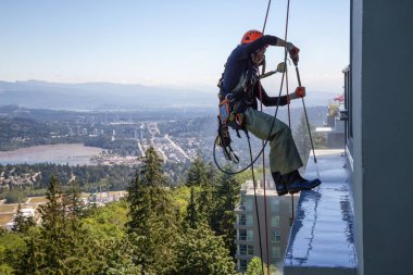 Burnaby Mountain, Vancouver, British Columbia, Canada - July 011, 2018: High rise rope access window cleaner is power washing the building during a hot sunny summer day. clipart