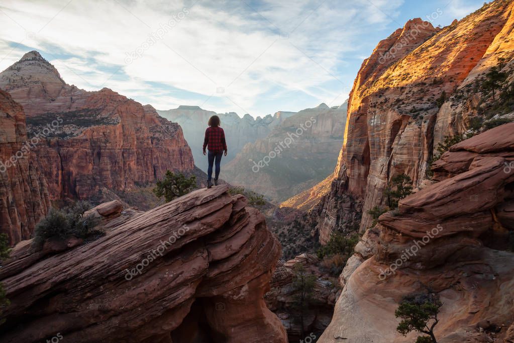 Adventurous Girl at the edge of a cliff is looking at a beautiful landscape view in the Canyon during a vibrant sunset. Taken in Zion National Park, Utah, United States.