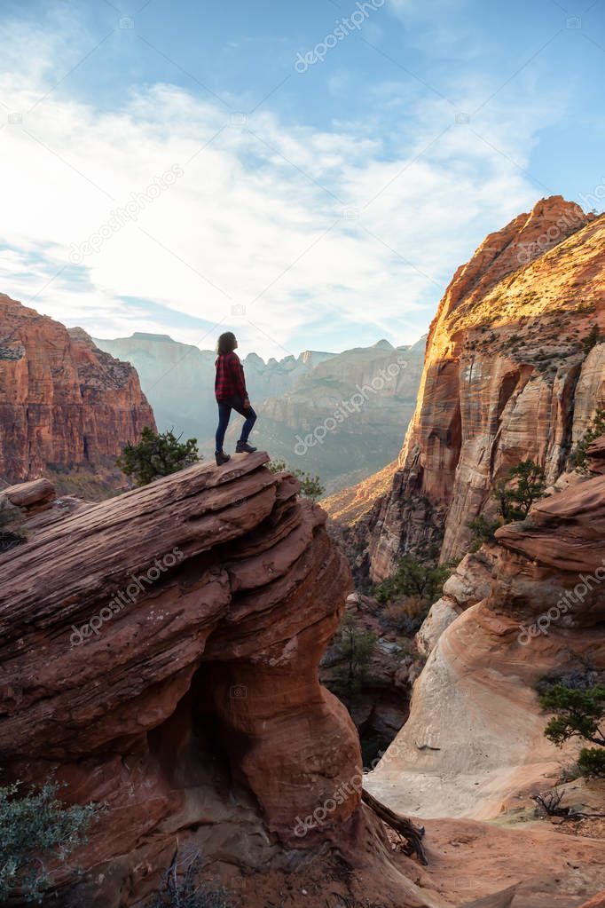 Adventurous Girl at the edge of a cliff is looking at a beautiful landscape view in the Canyon during a vibrant sunset. Taken in Zion National Park, Utah, United States.