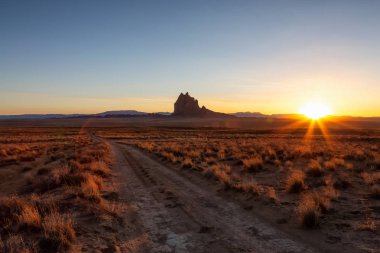Striking landscape view of a dirt road in the dry desert with a mountain peak in the background during a vibrant sunset. Taken at Shiprock, New Mexico, United States. clipart