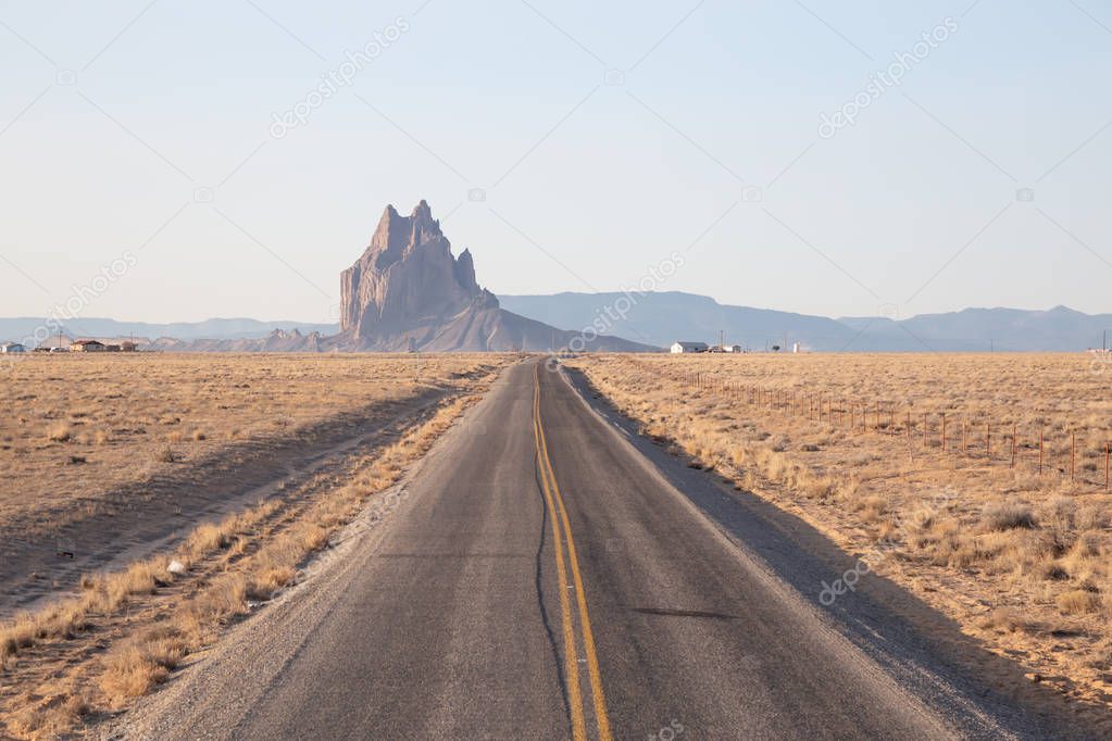 View of a road in a dry desert with a Shiprock mountain peak in the background during a vibrant sunny sunrise.Taken at Rattlesnake, New Mexico, United States.
