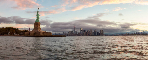 Panoramic view of the Statue of Liberty and Downtown Manhattan in the background during a vibrant cloudy sunrise. Taken in Jersey City, New Jersey, United States.