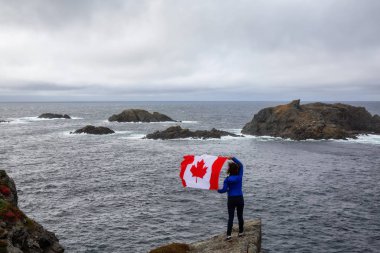 Adventurous woman holding a Canadian Flag on a Rocky Atlantic Ocean Coast during a cloudy day. Taken in Sleepy Cove, Crow Head, Twillingate, Newfoundland, Canada. clipart