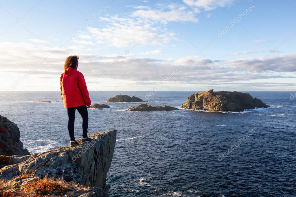 Woman in red jacket is standing at the edge of a cliff and enjoying the beautiful ocean scenery. Taken in Crow Head, North Twillingate Island, Newfoundland and Labrador, Canada.