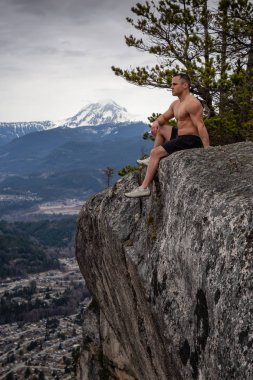 Fit ve Kaslı Kafkas Adam bir dağın tepesindeki kayalıkta oturuyor ve manzaranın tadını çıkarıyor. Squamish, Kuzey Vancouver, Bc, Kanada'da çekildi.
