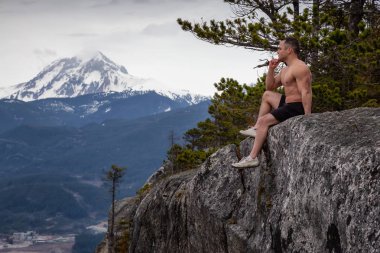 Fit ve Kaslı Kafkas Adam bir dağın tepesindeki kayalıkta oturuyor ve manzaranın tadını çıkarıyor. Squamish, Kuzey Vancouver, Bc, Kanada'da çekildi.