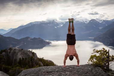 Fit and Muscular Young Man is doing a handstand on top of the Mountain during a cloudy day. Taken on Chief Mountain in Squamish, North of Vancouver, BC, Canada. clipart